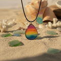 Rainbow sunset teardrop resin necklace on a black cord, displayed on sand with seashells and sea glass